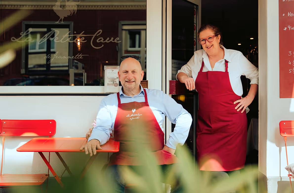 Ein lächelndes Paar in roten Schürzen vor dem Bistro La Petite Cave de Jeannette, mit roten Stühlen und Tischen.