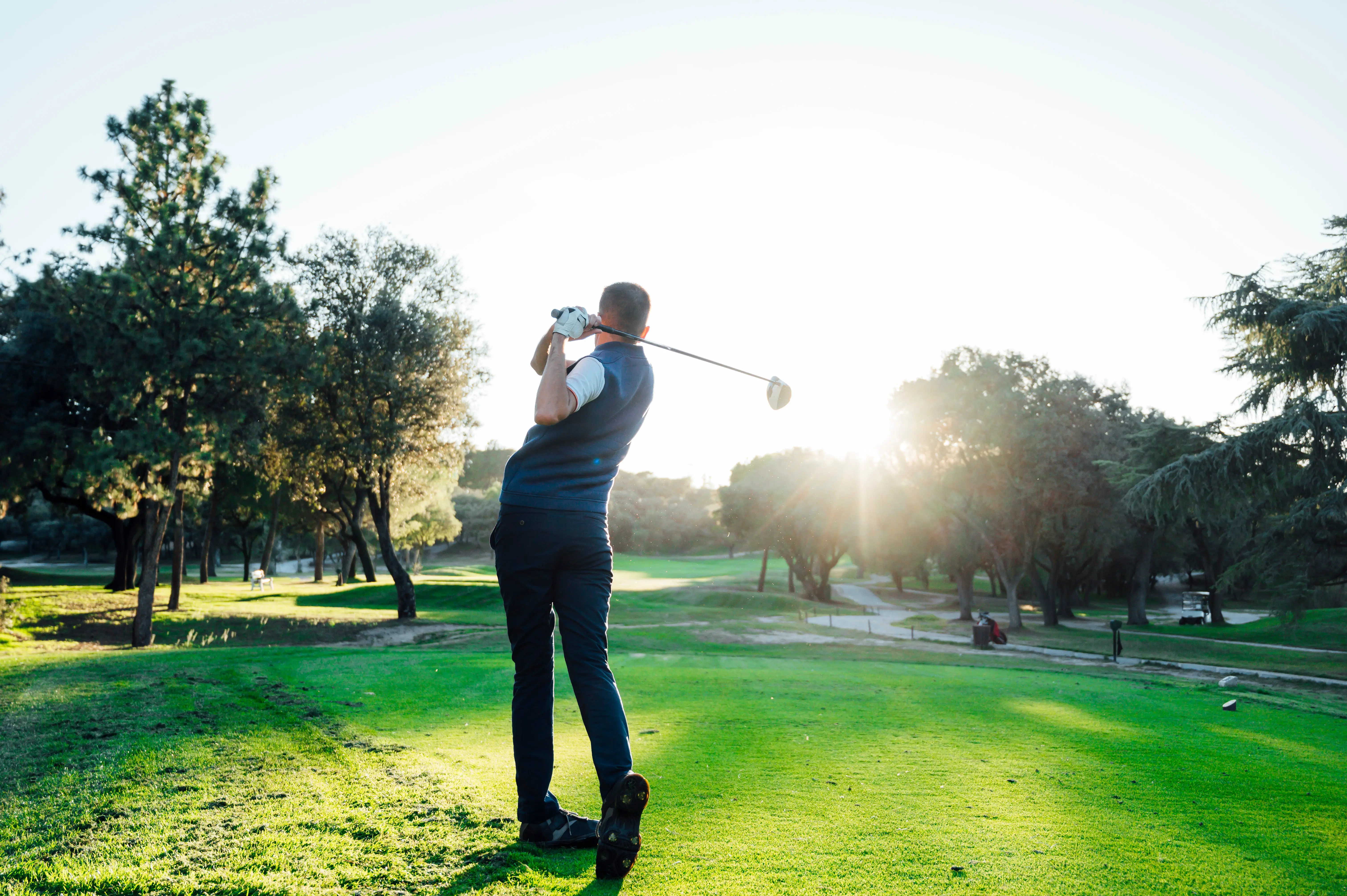 Golfer in mid-swing on a green golf course with trees and setting sun in the background.