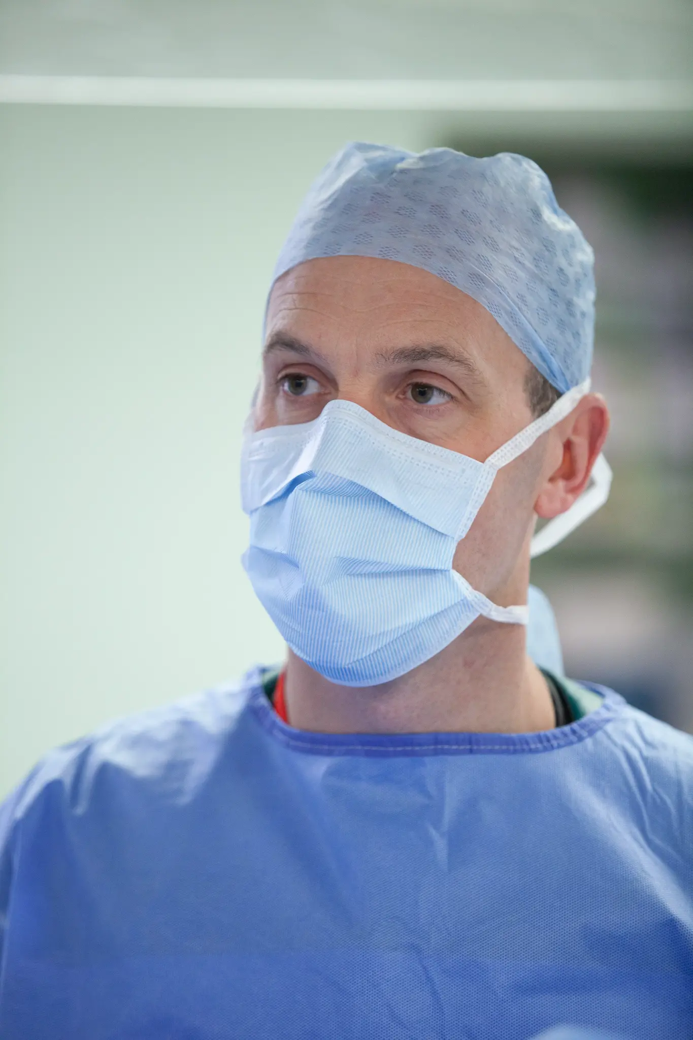 Male healthcare professional wearing a blue surgical cap, mask, and gown in a clinical setting.