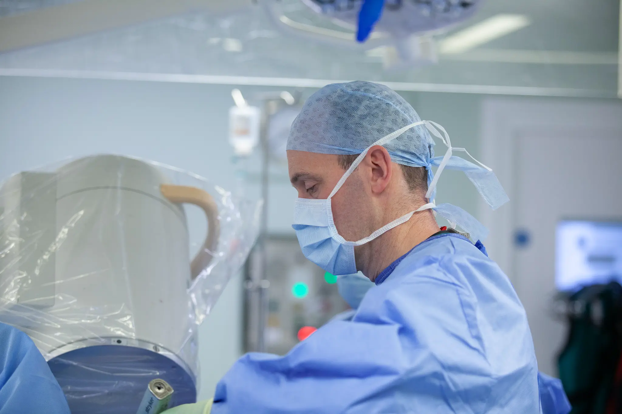 Surgeon in blue scrubs and surgical cap wearing a face mask, focused on a medical procedure in a hospital operating room.