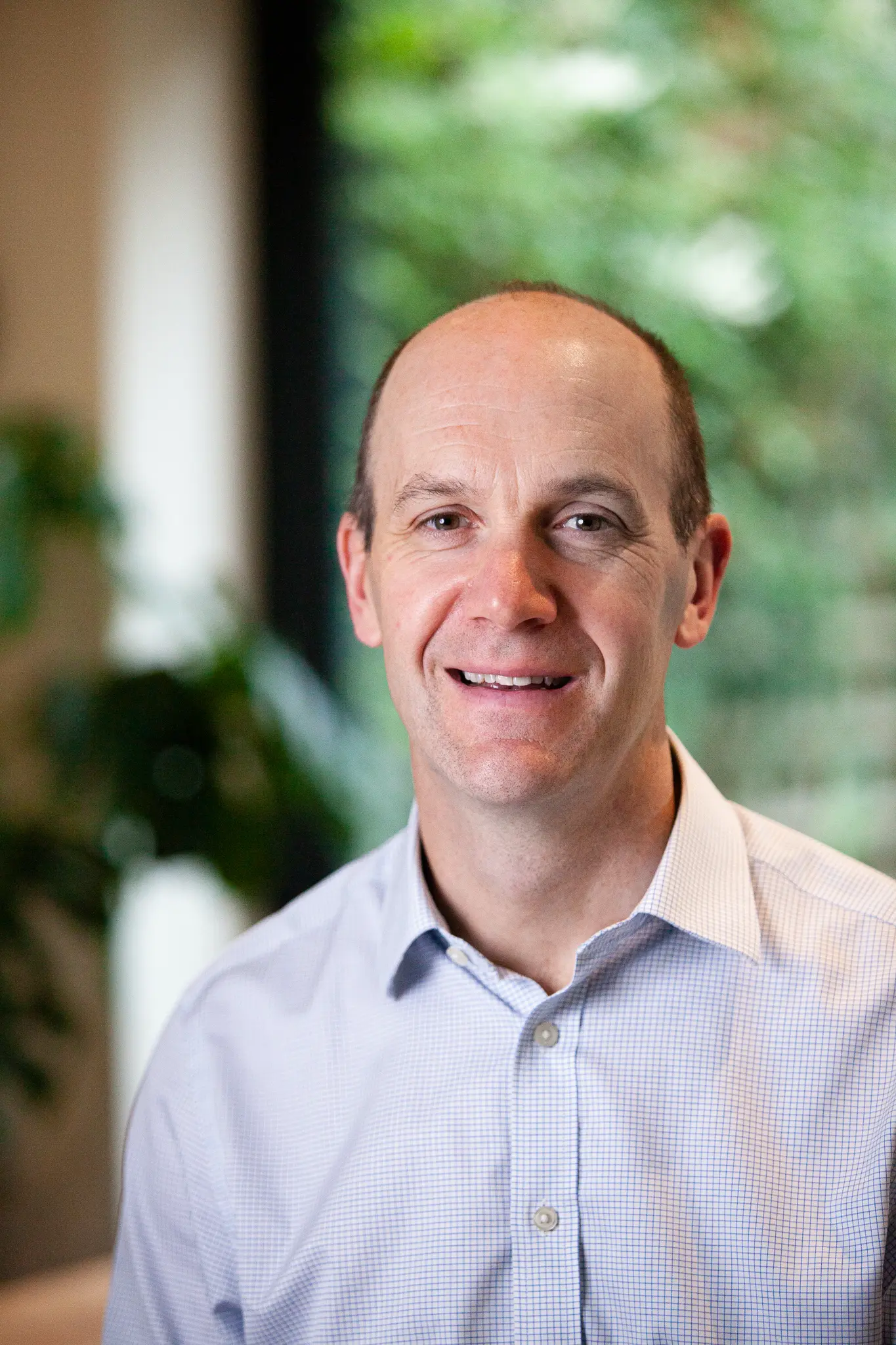 Smiling man with light skin and short hair wearing a light blue checkered shirt, with a blurred green background.