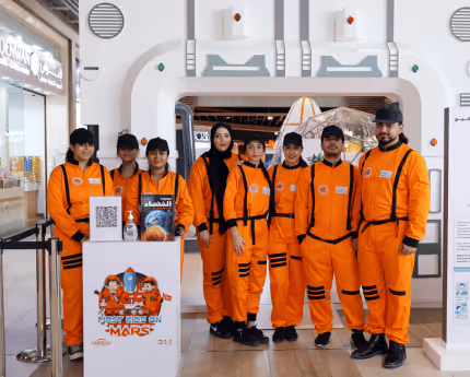 Group of people wearing bright orange astronaut suits posing together inside a space-themed exhibition area.