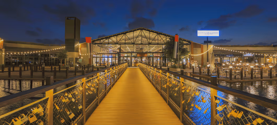 Golden-lit walkway leading to a modern glass building at night, reflected in the surrounding water.