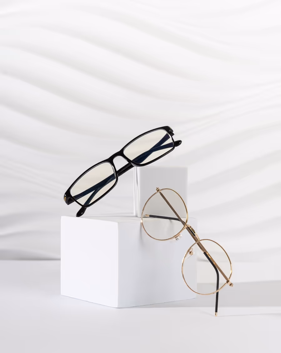 Two pairs of eyeglasses displayed on white geometric stands against a textured white background, one rectangular black frame and one round gold frame.