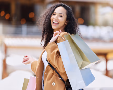 Smiling woman with curly hair carrying multiple shopping bags while walking outdoors.