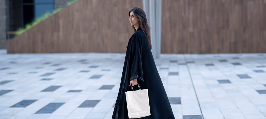 Woman in a black abaya and cap carrying a beige shopping bag while walking outdoors on a tiled pathway.