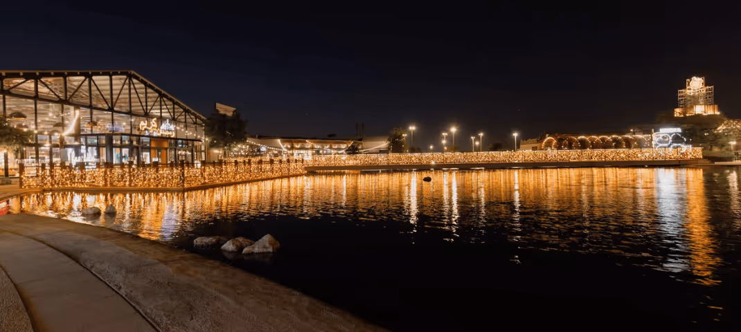 Al Khawaneej Walk at night, illuminated by festive lights reflecting across the waterfront and surrounding restaurants and walkways.