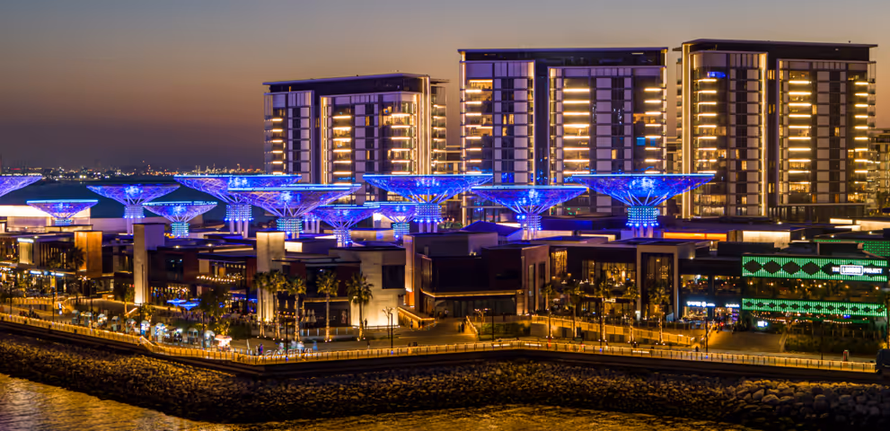 Night view of a modern waterfront district with illuminated blue canopies, lined with restaurants and shops, set against tall residential towers glowing with warm lights