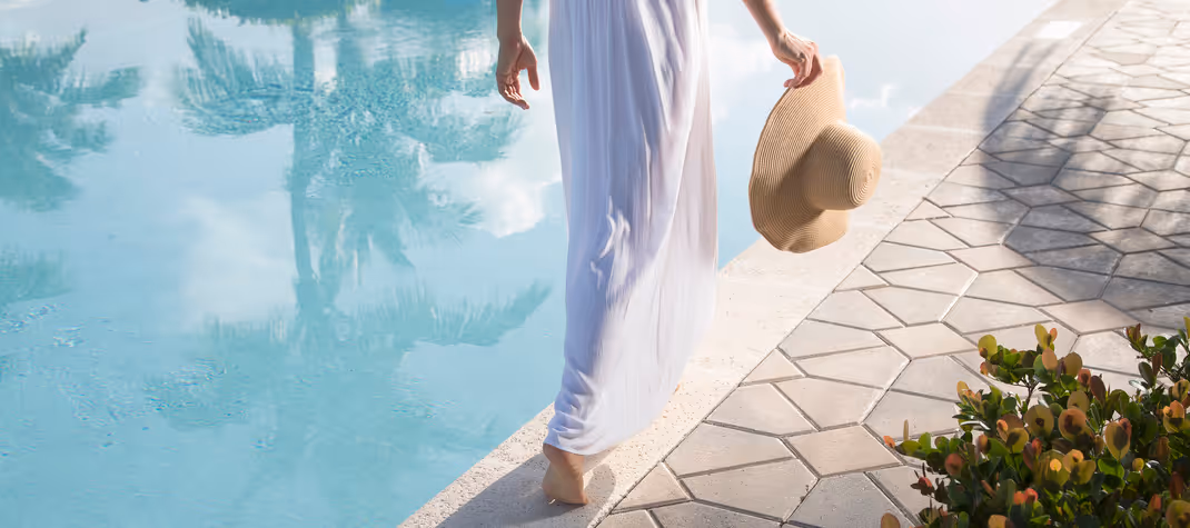 Woman walking barefoot beside a swimming pool, holding a sun hat, with palm tree reflections in the water.