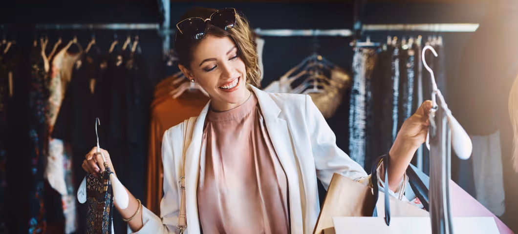 Smiling woman shopping for clothes, holding hangers with outfits while browsing through a clothing rack in a boutique.