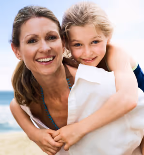 Smiling woman carrying a young child on her back at the beach with the sea and sky in the background.