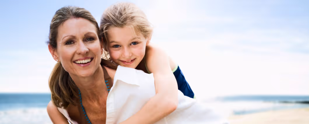Smiling woman carrying a young child on her back at the beach with the sea and sky in the background.