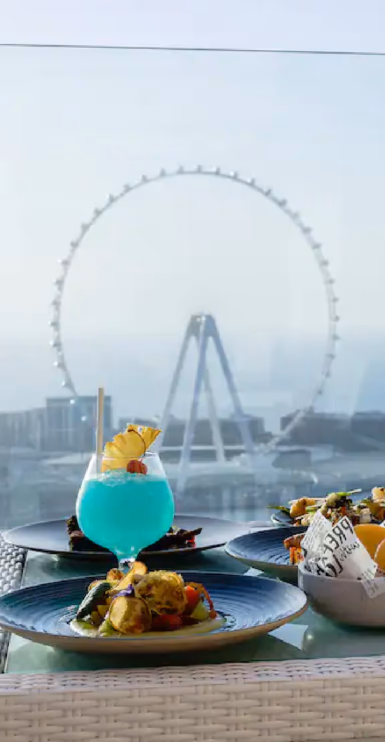 Plate of seafood and a vibrant blue cocktail on a table overlooking the sea with the Ain Dubai observation wheel in the background.