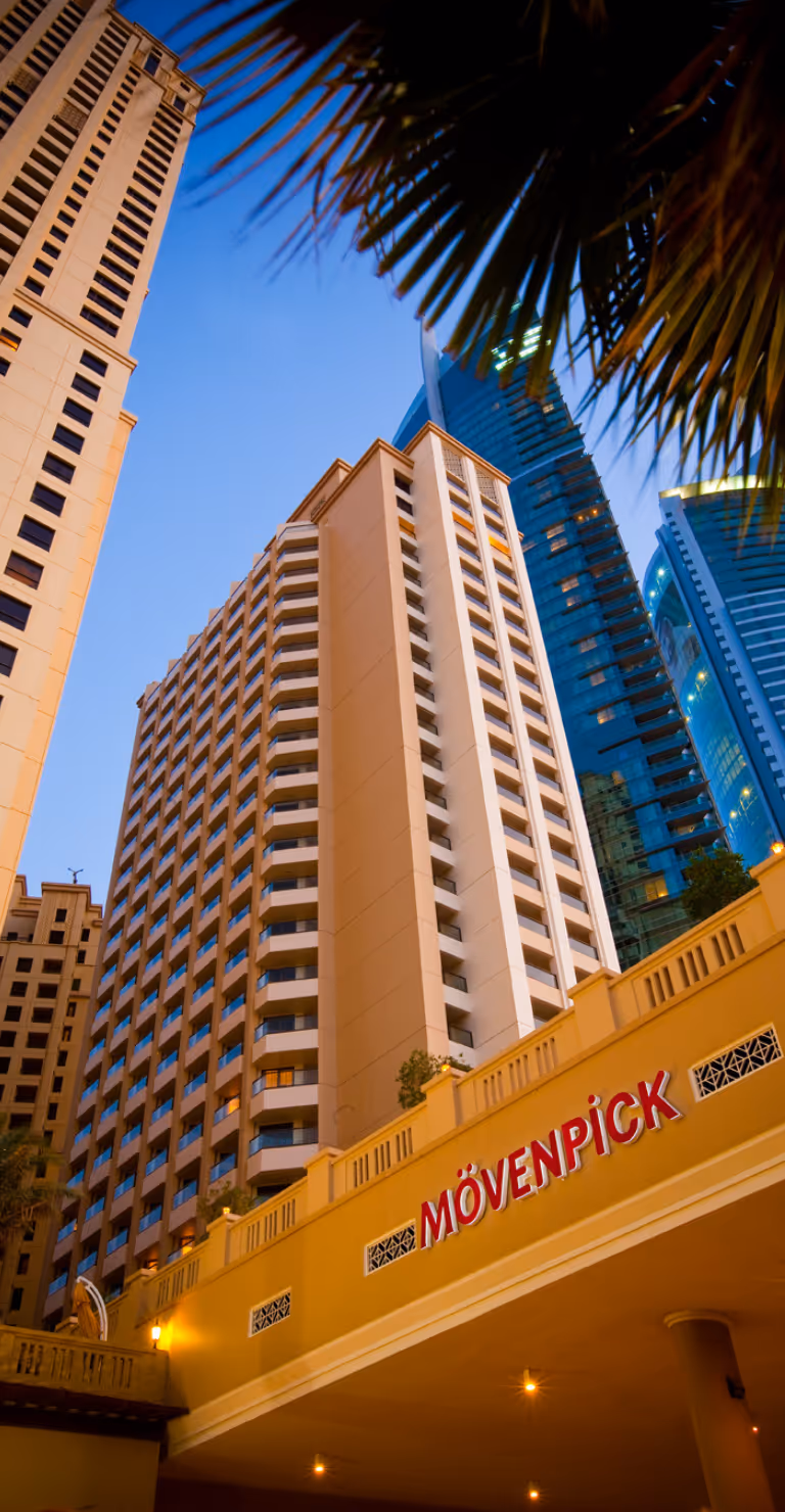 Low-angle view of tall beige hotel building with a pink illuminated 'Mövenpick' sign at the entrance, framed by modern skyscrapers and a palm frond against a clear blue sky.