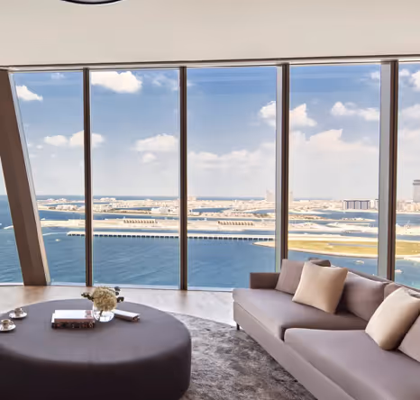 Modern living room with floor-to-ceiling windows offering a panoramic view of the sea and coastline, furnished with a sleek sofa, round coffee table, and neutral decor.