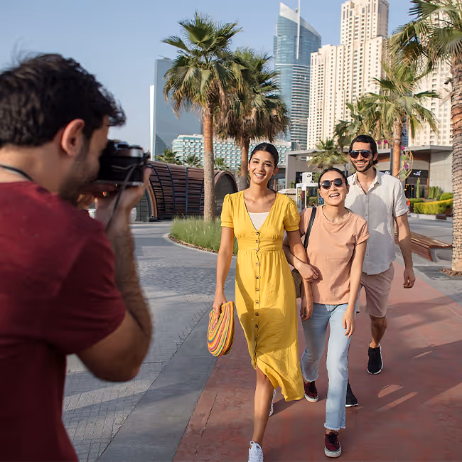 Photographer taking a picture of three smiling people walking outdoors on a palm-lined walkway with tall modern buildings in the background.