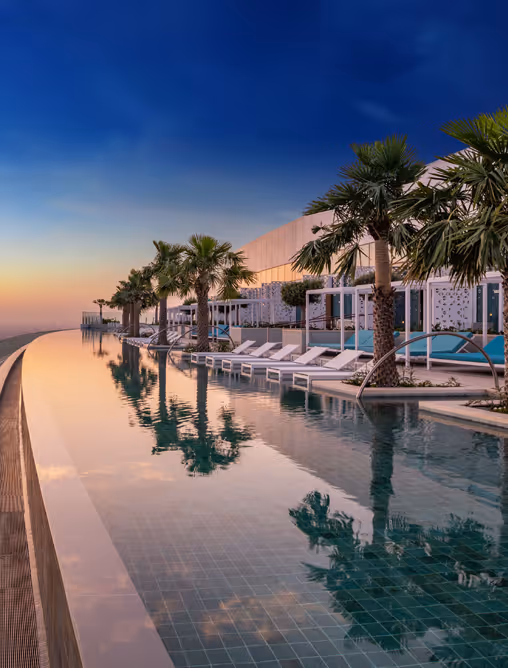 Infinity pool at sunset with lounge chairs, palm trees, and modern cabanas overlooking the ocean.