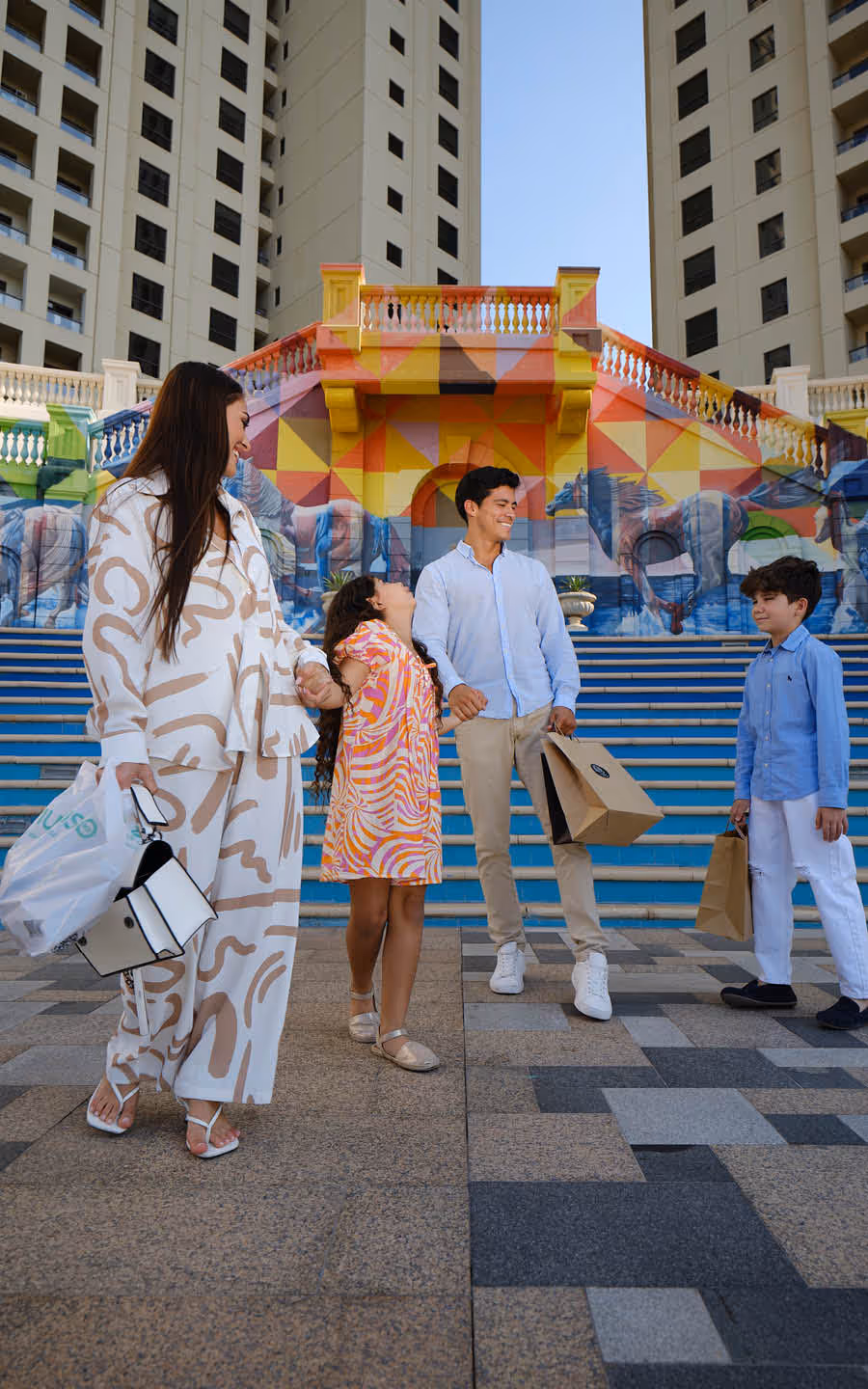 A family of four smiling and holding shopping bags while standing in front of colorful mural-covered stairs between tall buildings.