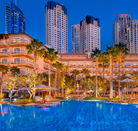 A large outdoor swimming pool surrounded by palm trees and a resort building, with tall city skyscrapers illuminated in the background at dusk.