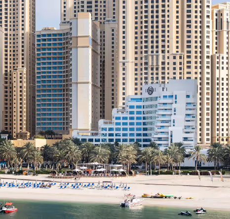 Beachfront hotel with white modern architecture, surrounded by tall skyscrapers, palm trees, and sun loungers along the sandy shore.