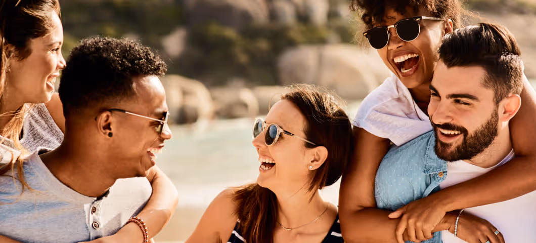 A group of friends enjoying a sunny day at the beach, smiling and laughing together. Some are wearing sunglasses, and the mood is cheerful and relaxed.