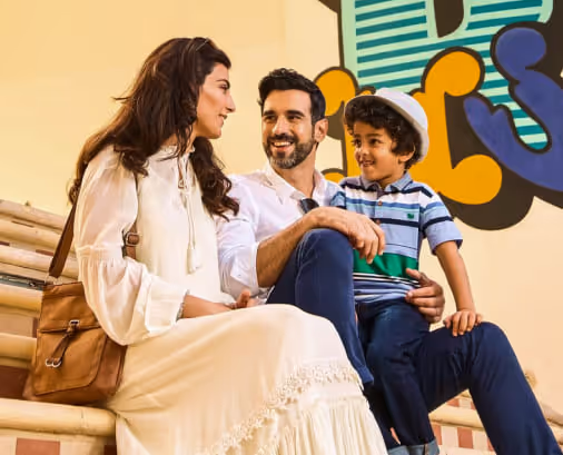 A family sitting together on tiled steps, smiling and talking. The mother wears a long white dress, the father is dressed casually in white and navy, and their young son in a striped shirt and hat sits on his father’s lap. A colorful wall mural is in the background.