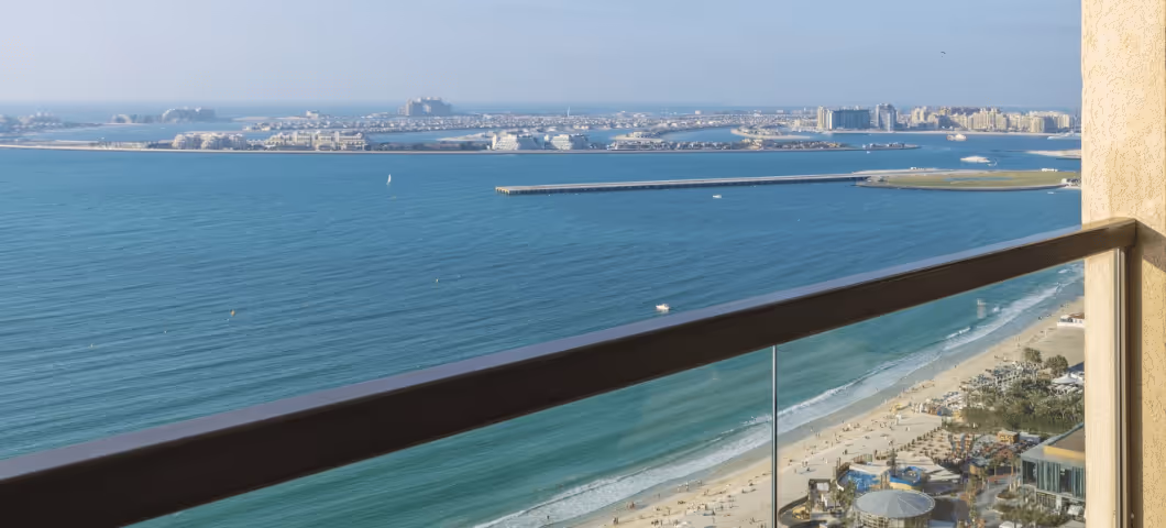 High-rise balcony view overlooking a sandy beach, turquoise sea, and the man-made Palm Jumeirah islands in the distance.