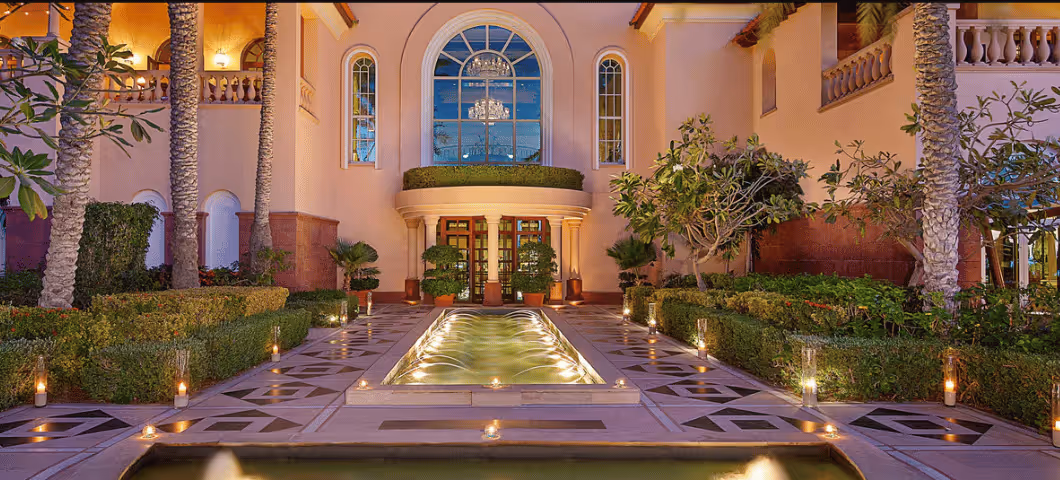 Elegant courtyard with a central water feature, trimmed hedges, and candlelit pathways leading to an arched entrance with a large decorative window.
