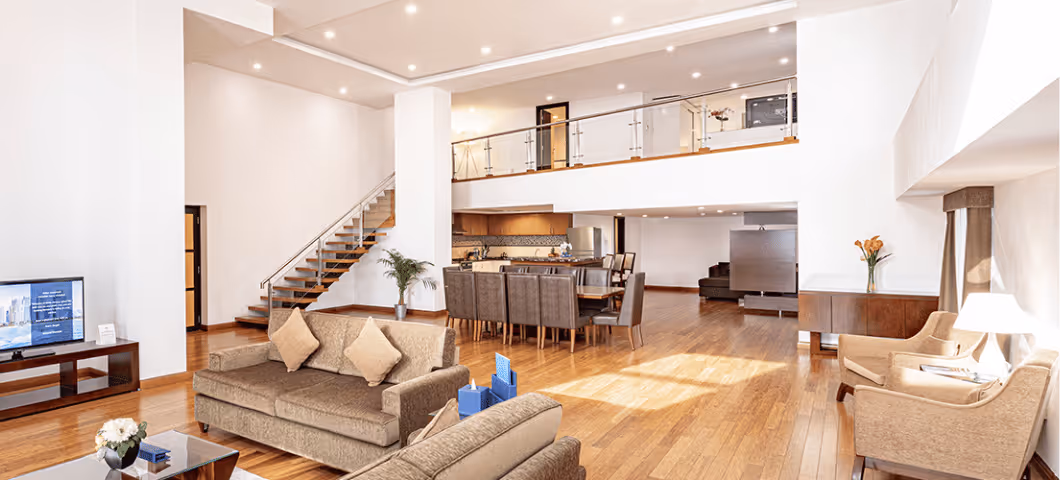 Spacious modern living room with brown sofas, wooden flooring, open kitchen, and an upper mezzanine level.