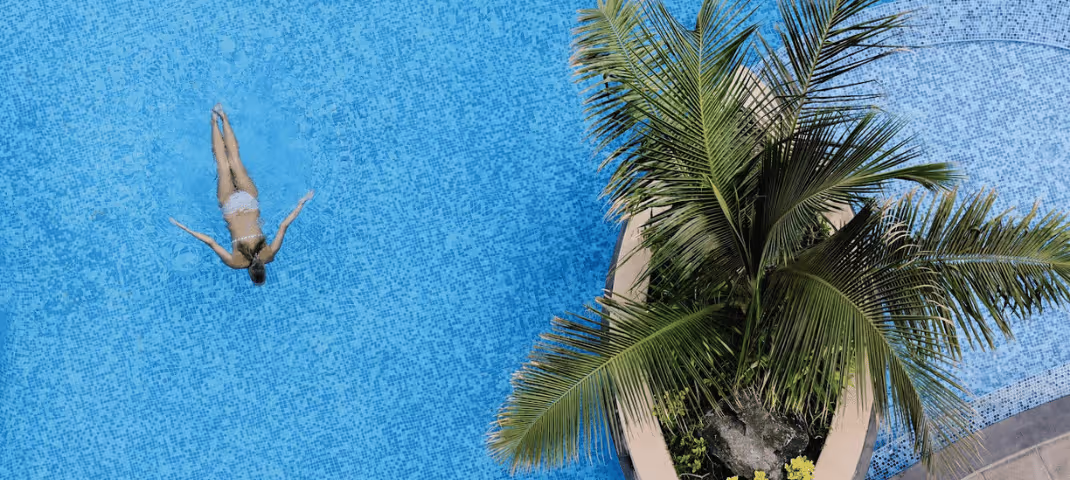 A person floating on their back in a bright blue swimming pool next to tropical palm plants.