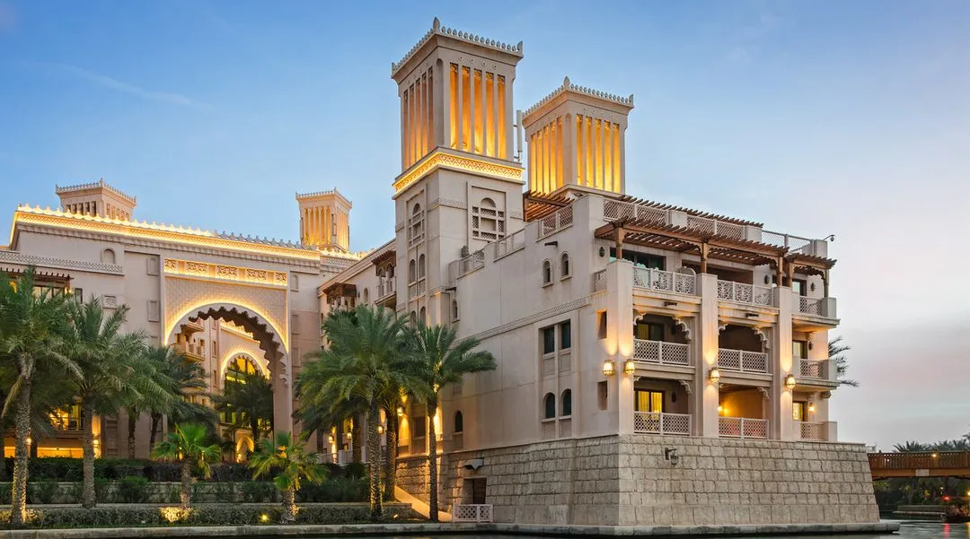 Luxurious Middle Eastern-style hotel building with wind towers, intricate archways, and warm golden lighting surrounded by palm trees at sunset.