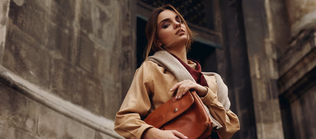 Fashionable woman in a beige trench coat holding a brown leather handbag, standing against an old stone building backdrop.