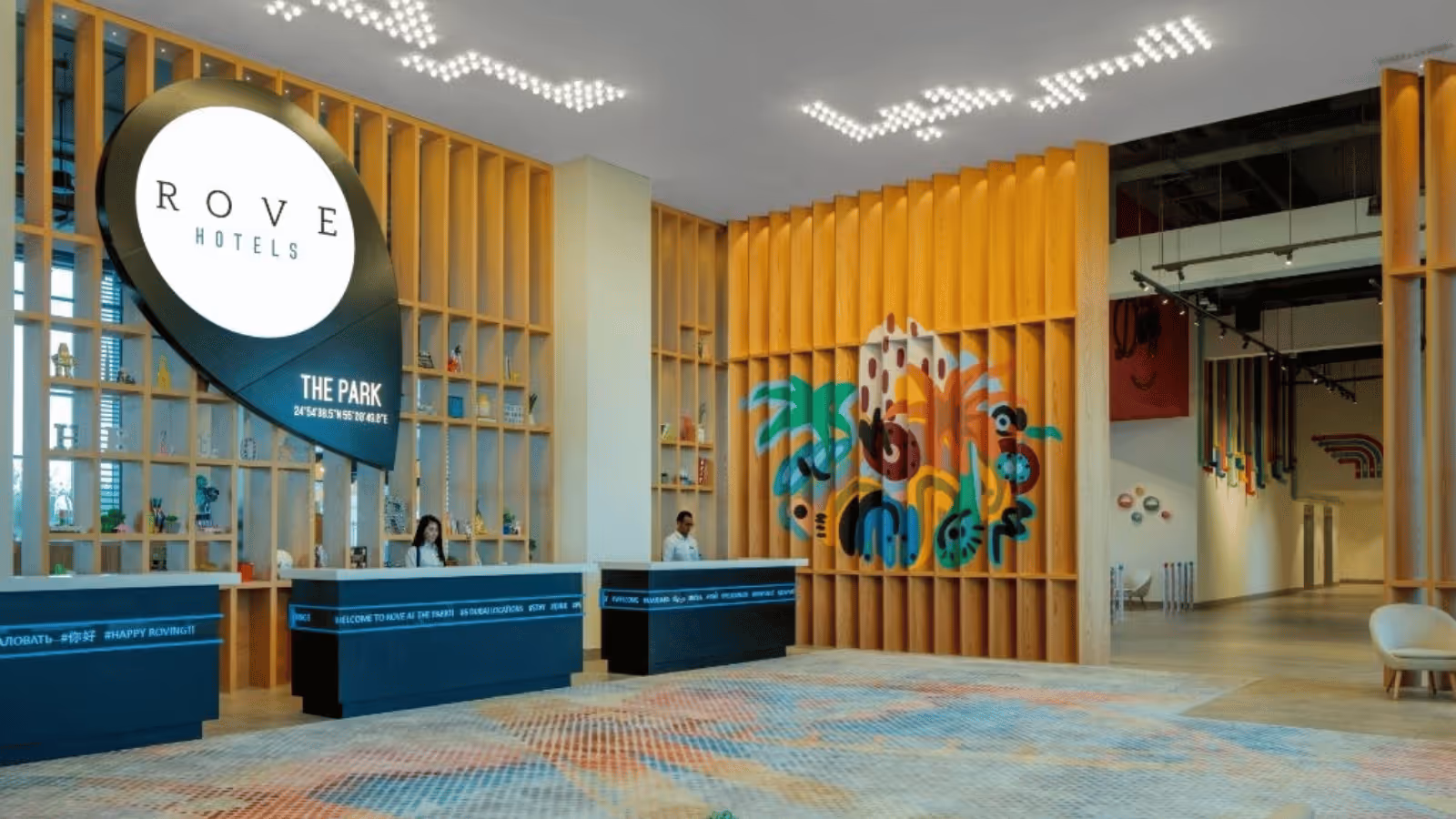Modern hotel lobby of Rove Hotels with blue reception desks, wooden partitions, colorful wall art, and geometric ceiling lights.
