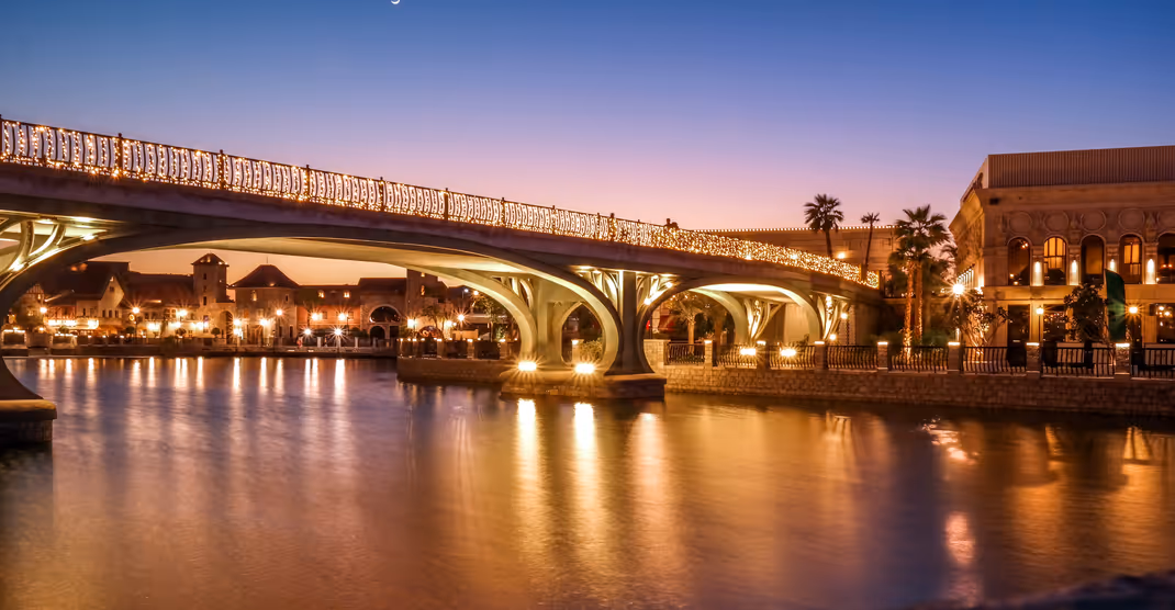 Illuminated bridge over a calm river at dusk, with reflections of golden lights shimmering on the water and surrounding buildings softly lit in the evening glow.