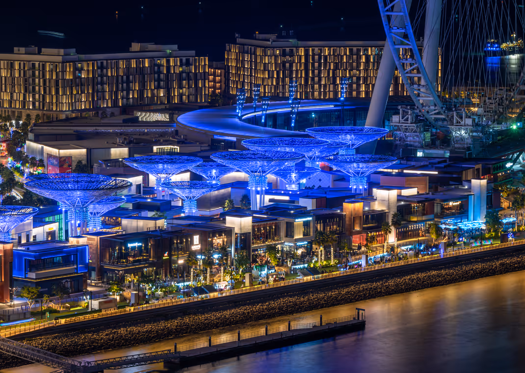 Vibrant night view of Bluewaters Island in Dubai, showcasing a lively dining and entertainment area illuminated with blue lighting, with Ain Dubai observation wheel in the background.