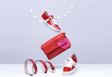 Floating fashion accessories display with red sneakers, a red handbag with a pink accent, and three red leather bracelets against a light background.