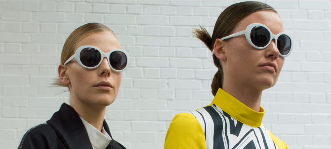 Two people wearing large round white sunglasses and stylish outfits with bold patterns standing against a light brick wall.
