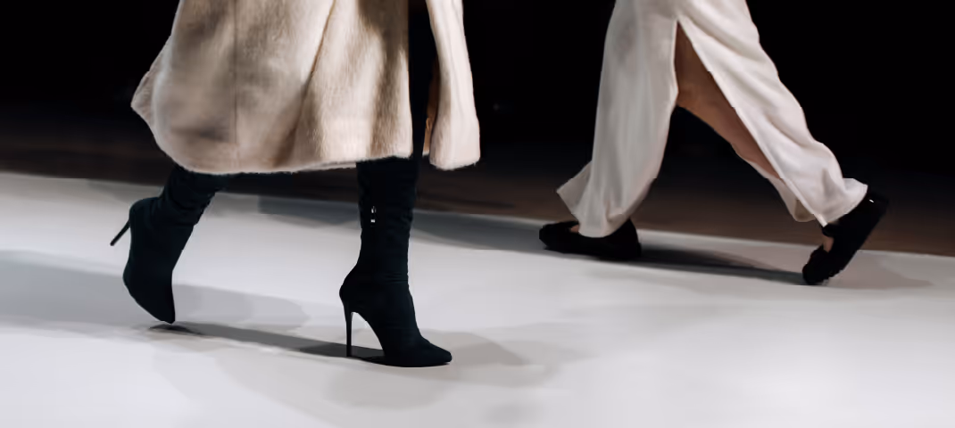 Two models walking on a runway, wearing neutral-toned outfits with long coats and heeled boots.

