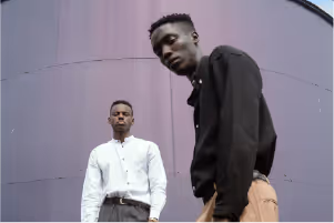 Two people posing outdoors against a large purple wall, one wearing a white shirt and the other in a black shirt with tan pants.
