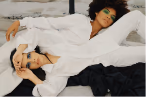 Two people dressed in white outfits lying on a light surface, posing with relaxed expressions and artistic green eye makeup.
