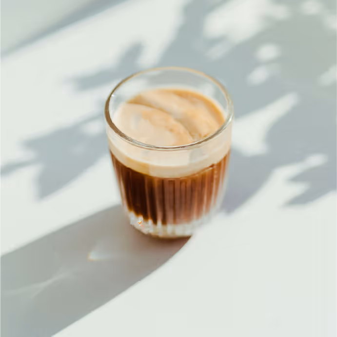 Glass of coffee with milk foam on a white surface, with soft natural light and leaf shadows.

