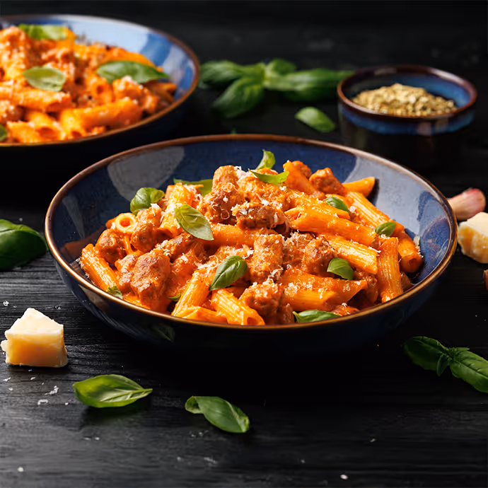Bowl of penne pasta with tomato sauce, ground meat, and fresh basil leaves on a dark table.
