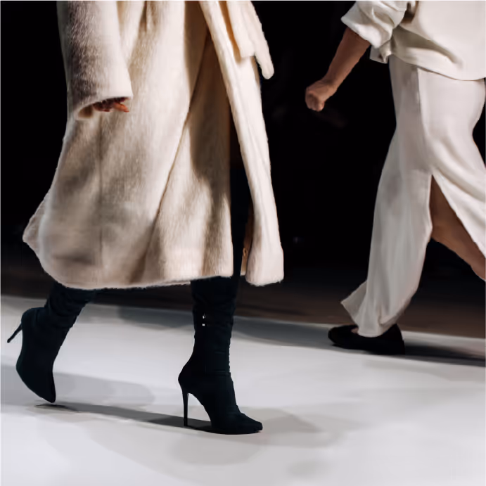 Two models walking on a runway, wearing neutral-toned outfits with long coats and heeled boots.
