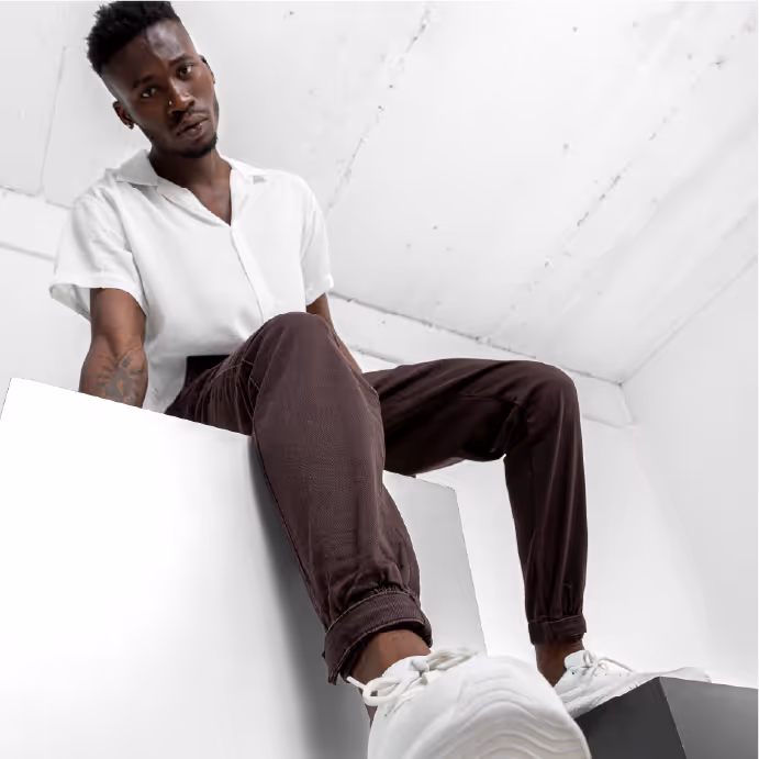Person sitting on a white platform in a minimal studio wearing a white shirt, dark pants, and white sneakers.
