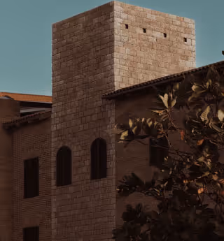 Stone building with arched windows partially obscured by tree branches, under a clear sky.

