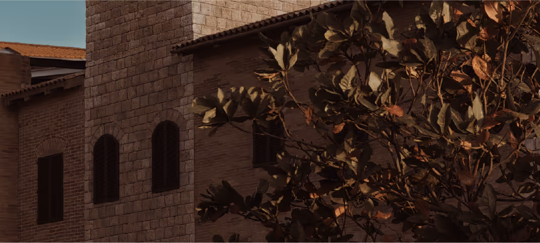 Stone building with arched windows partially obscured by tree branches, under a clear sky.
