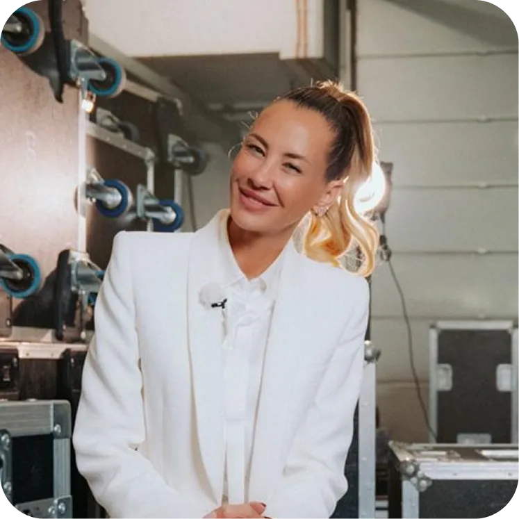 Lächelnde Frau mit hohem Pferdeschwanz trägt ein weißes Blazer-Outfit in einem Raum mit technischem Equipment.