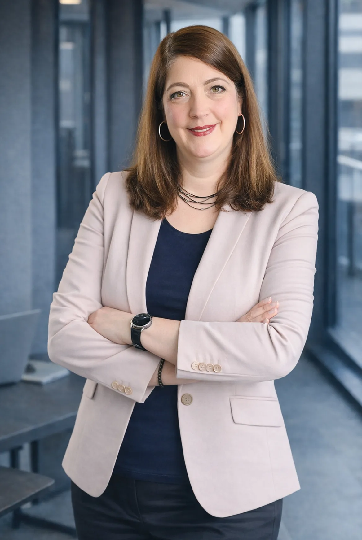 Frau mit rosa Blazer, blauem Hemd und verschränkten Armen vor Bürofenstern.