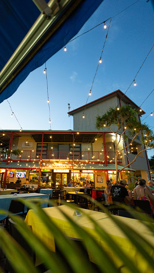 Outdoor dining area with string lights and people seated at tables during twilight in front of a two-story building.