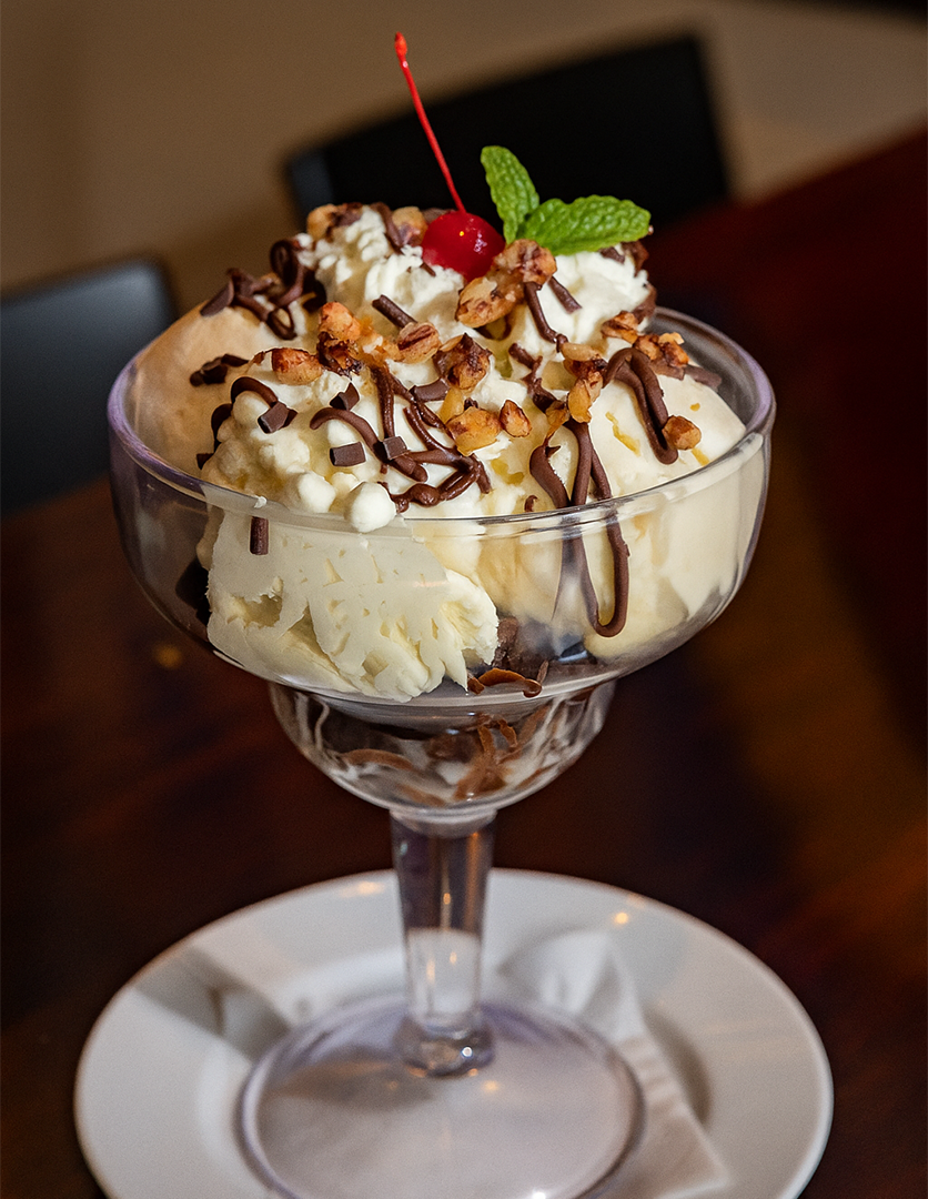 Ice cream sundae with whipped cream, chocolate drizzle, chopped nuts, a cherry, and a mint leaf in a glass dessert cup on a white plate.